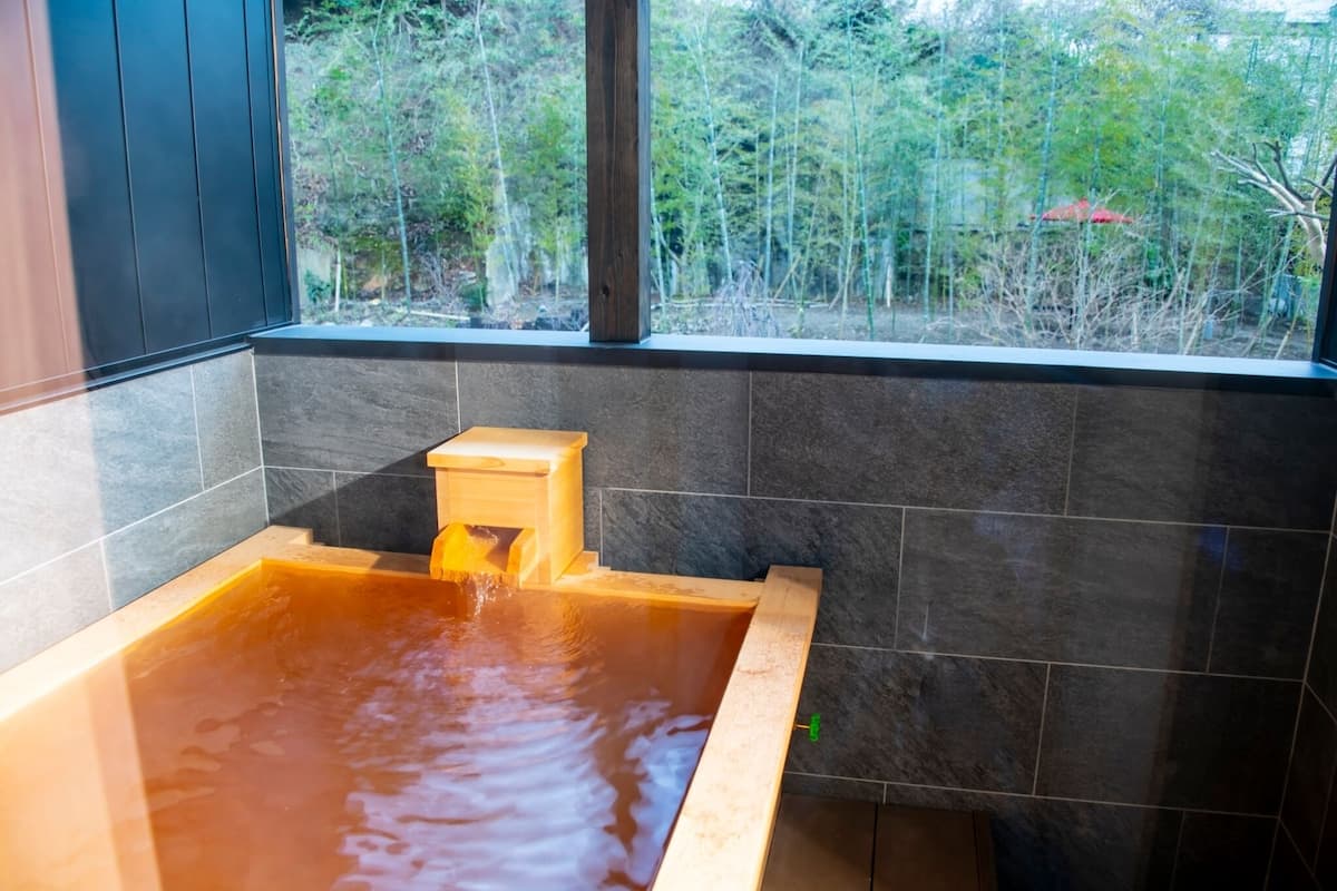 Ein Whirlpool mit Holzstufe, umgeben von dunklen Fliesenwänden und einem Fenster mit Blick auf ein bewaldetes Gebiet.