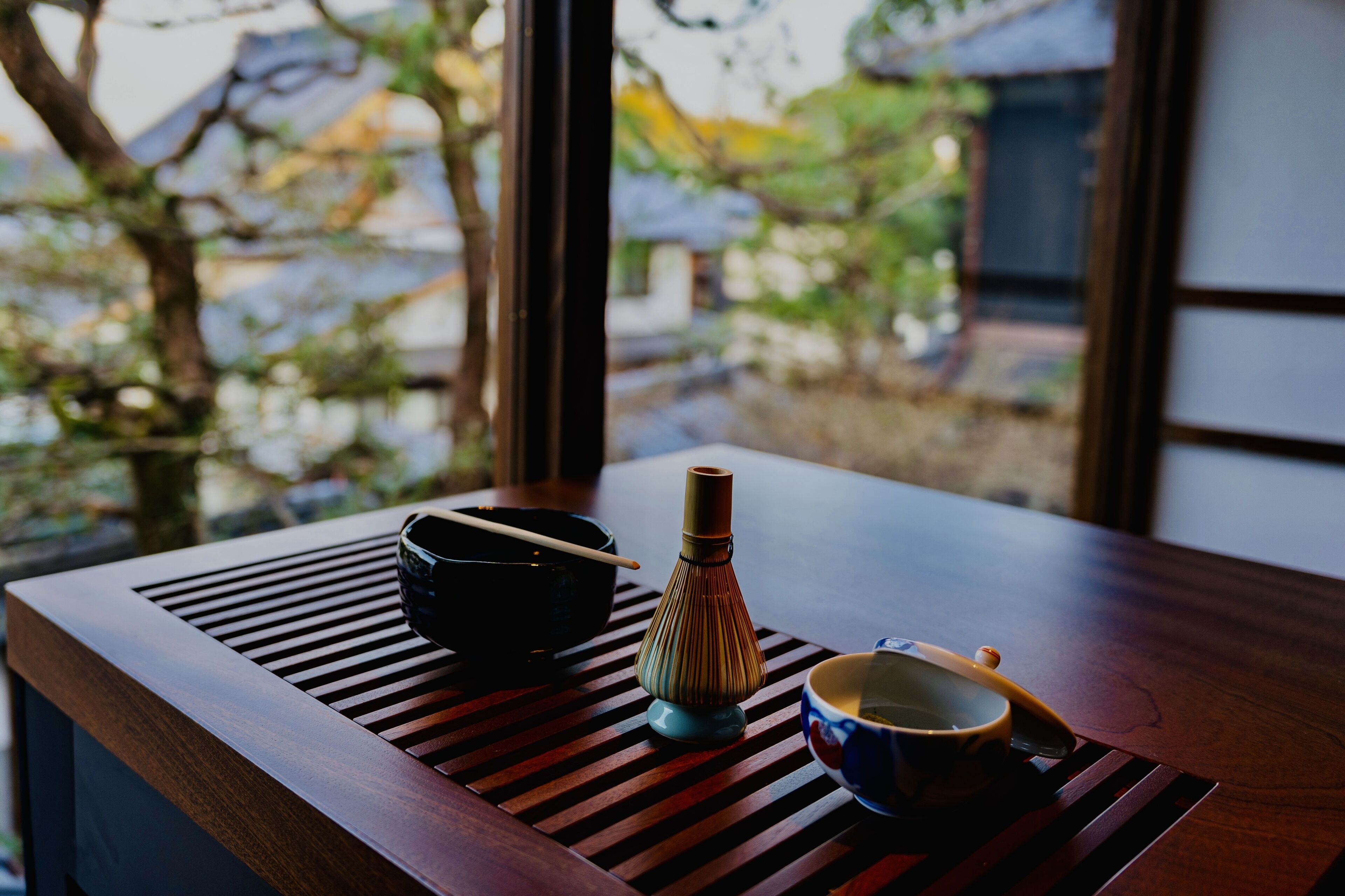 A wooden table with a black bowl, a traditional Japanese tea set, and a view of a traditional building through a window.