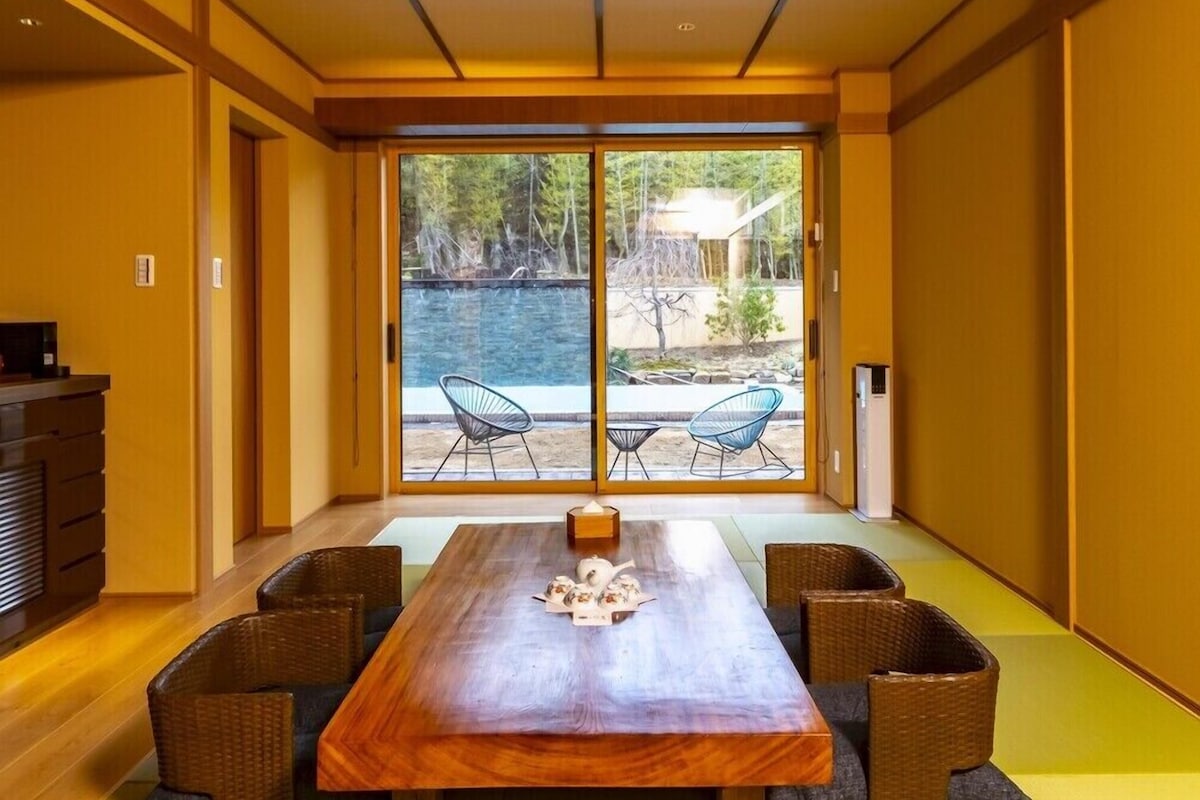 A dining area with a wooden table and chairs, a view of a natural landscape through large sliding doors.