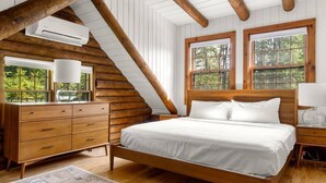 Un dormitorio de madera con cama, cómoda y dos ventanas que dan a zonas verdes.