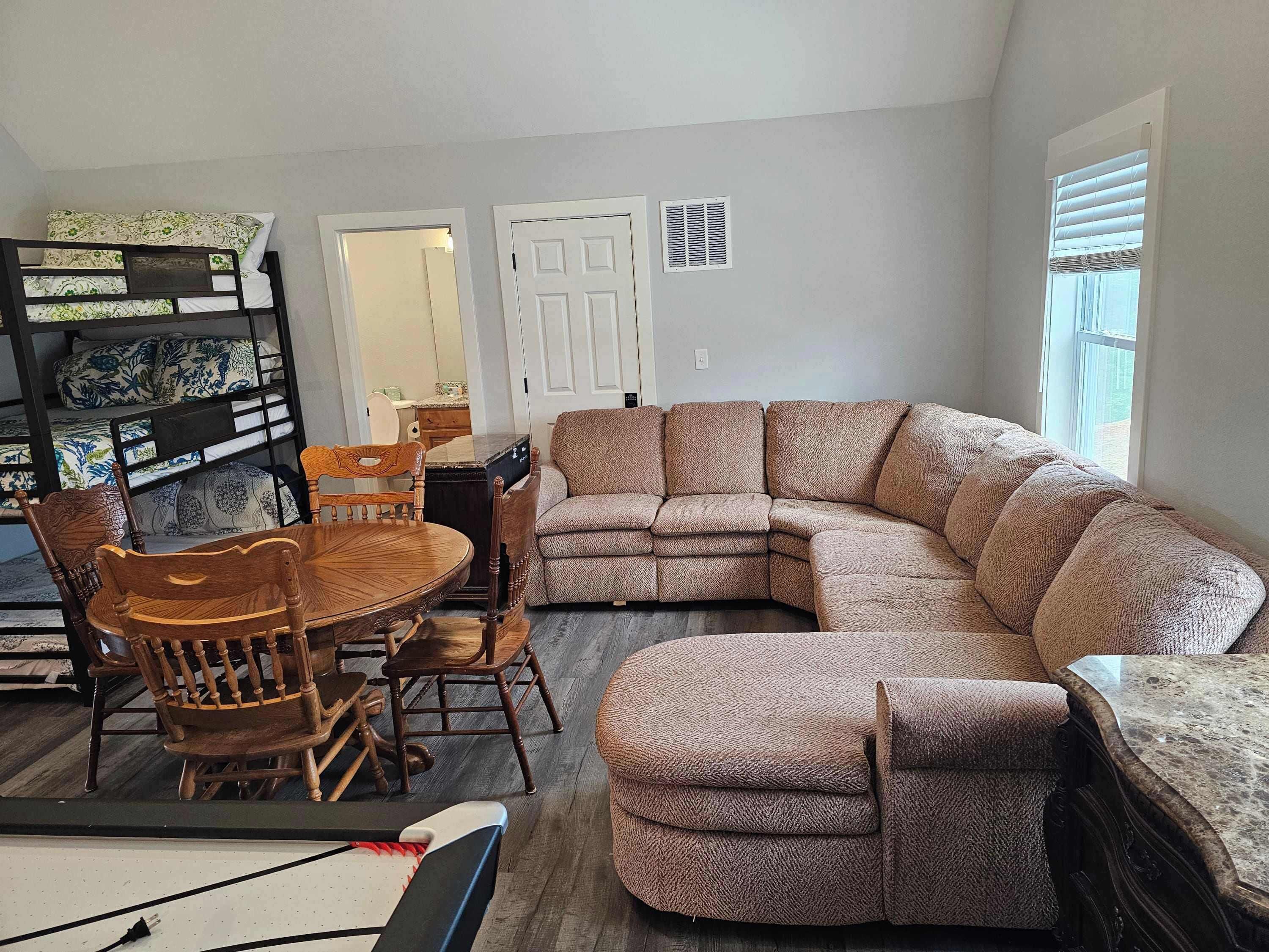 A living room with a sectional sofa, wooden chairs, a dining table, and a bunk bed.