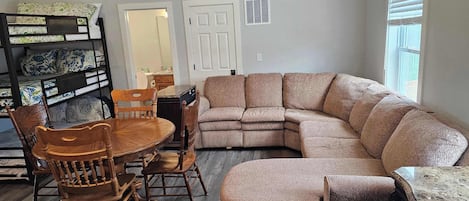 A living room with a sectional sofa, wooden chairs, a dining table, and a bunk bed.