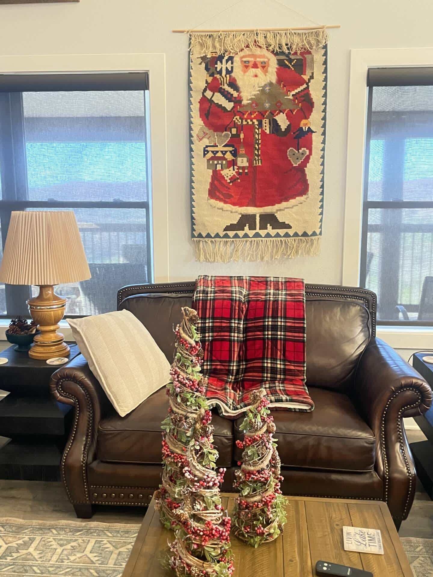 A living room with a brown leather sofa, a plaid blanket draped over it, a decorative wreath on a coffee table, and a wall tapestry.