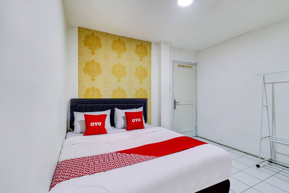 Habitación de hotel con una cama, dos almohadas rojas con el texto "OYO", una pared amarilla estampada y una puerta blanca.