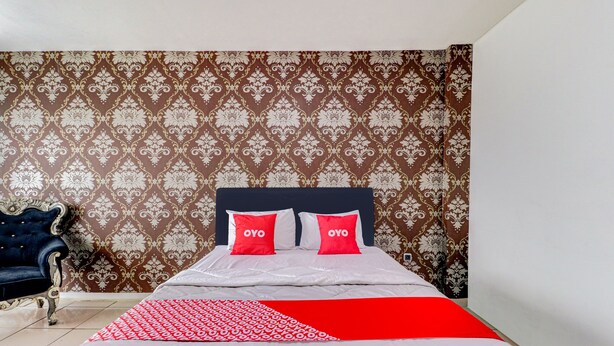 Un lit bien fait, agrémenté de deux oreillers rouges, d’un plaid rouge et blanc à motifs et d’une tête de lit bleu foncé. La pièce est décorée d’un papier peint à motifs et comprend un fauteuil bleu foncé.