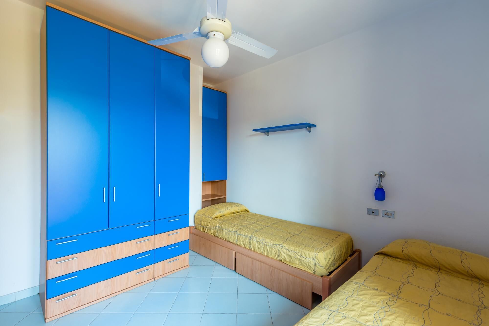 Une chambre à coucher comprenant un lit, une armoire, un ventilateur de plafond et une étagère fixée au mur.