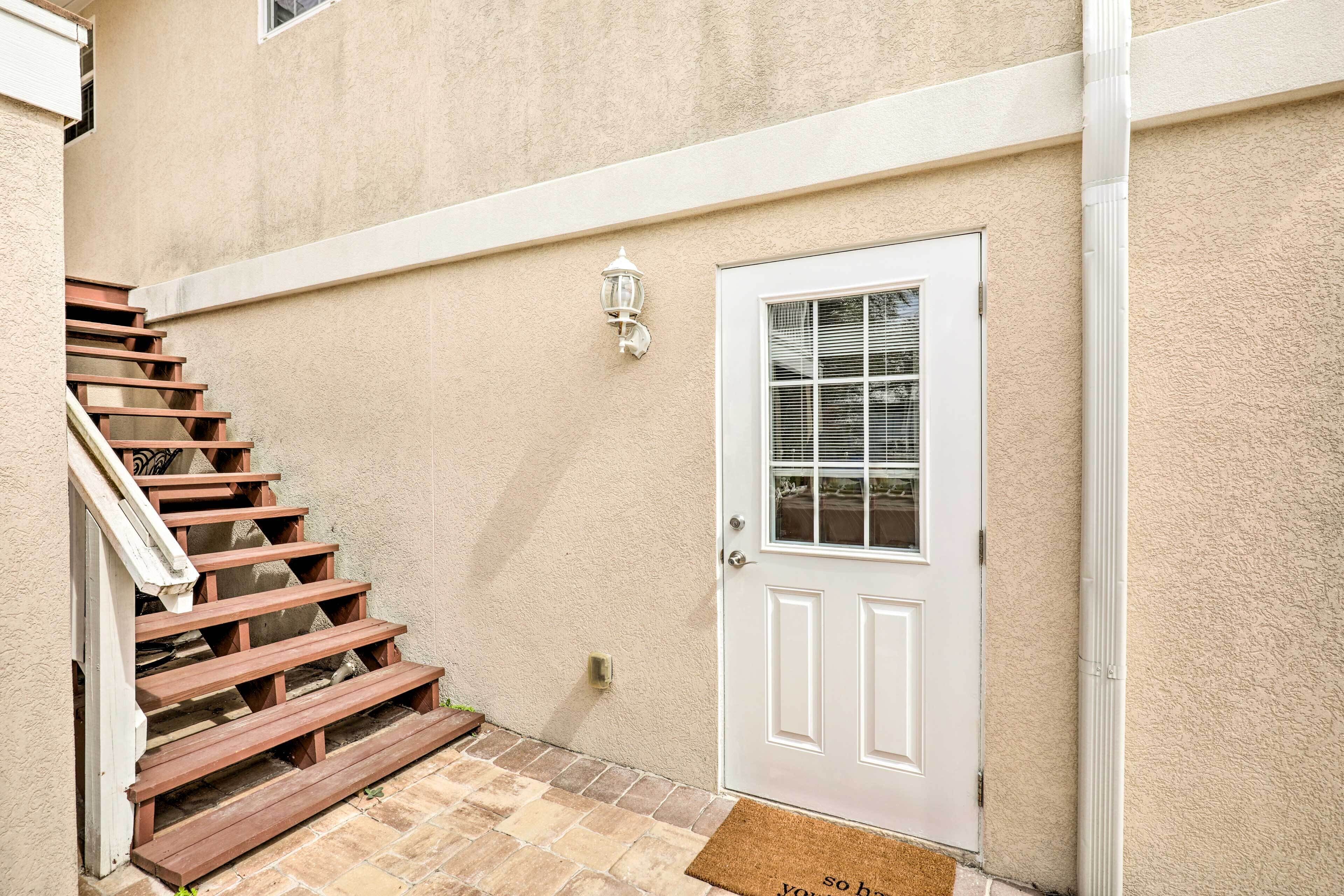 Una pared exterior color beige, con una puerta blanca y una escalera de madera que conduce a un porche.