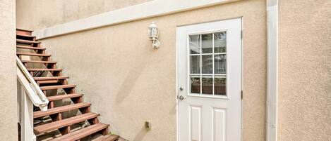 Una pared exterior color beige, con una puerta blanca y una escalera de madera que conduce a un porche.