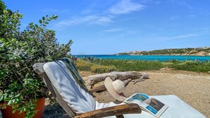 A beach chair with a book and a straw hat, overlooking a coastal landscape with clear blue waters and rocky hills.