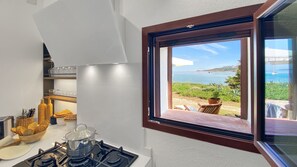 A kitchen with a gas stove, a window overlooking a scenic coastal view. - La Maranita Apartment (La Maddalena)