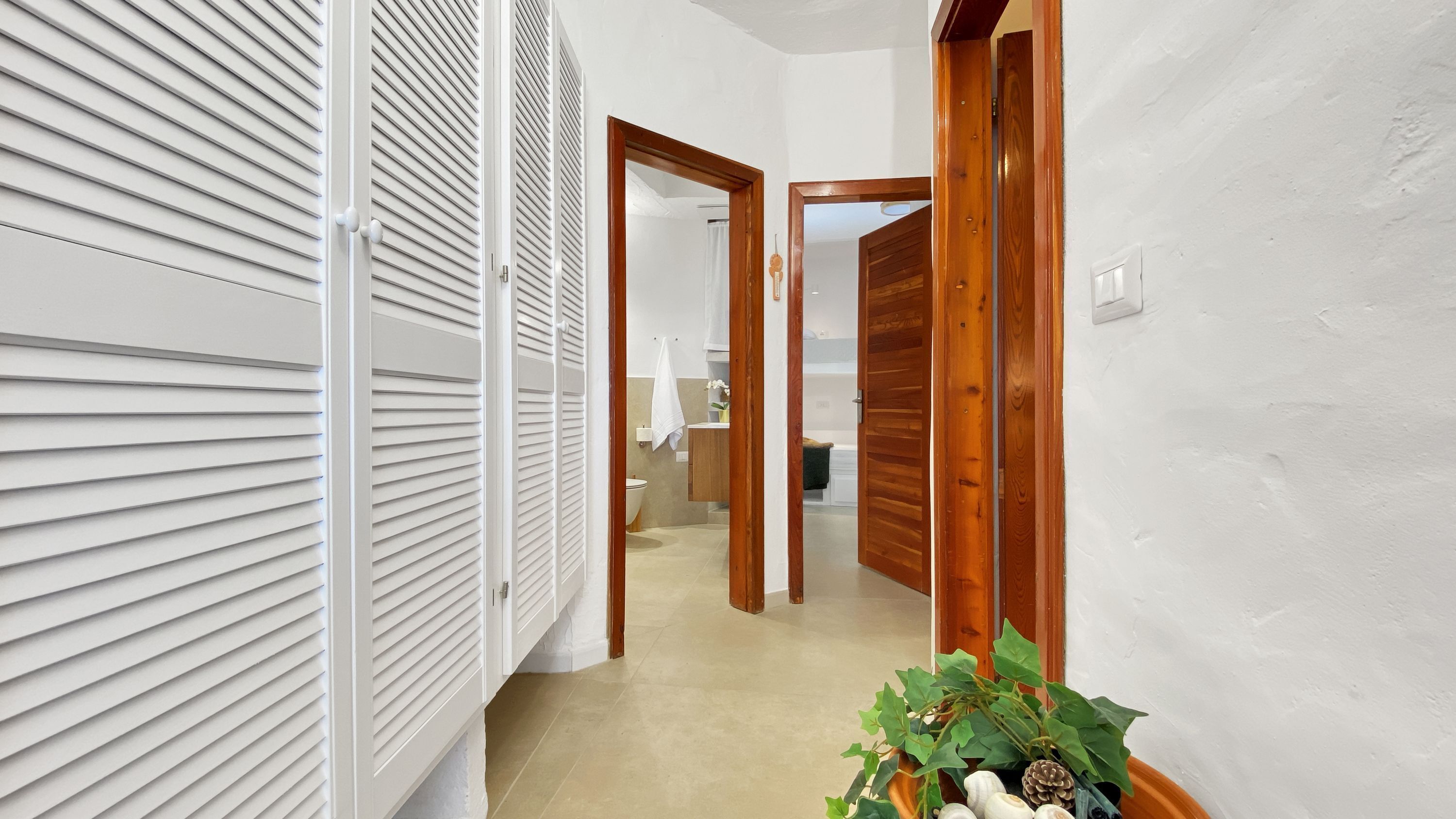A hallway with white blinds, wooden doors, and a plant with pinecones and greenery.
