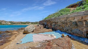 A rocky beach with a stone wall, a striped towel, and a hat. - La Maranita Apartment (La Maddalena)