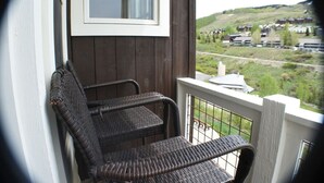 Een balkon met rieten stoelen, uitzicht op een stad en heuvels, en een raam boven de stoelen.