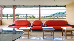 Un espace détente avec des canapés rouges et de petites tables, de grandes fenêtres offrant une vue sur des collines verdoyantes et un ciel dégagé.
