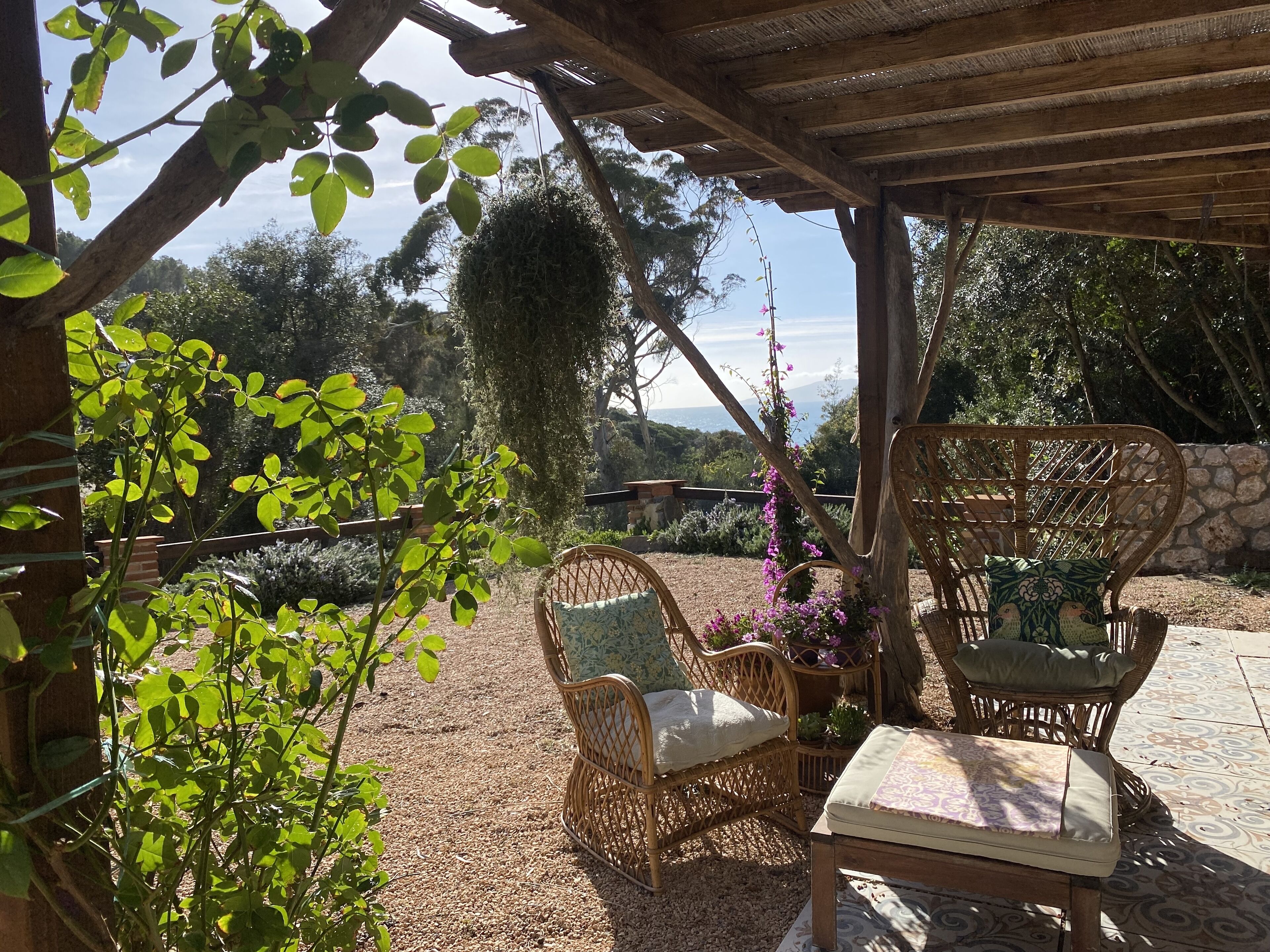 A covered outdoor seating area with wicker chairs and a table, surrounded by greenery and a stone wall.