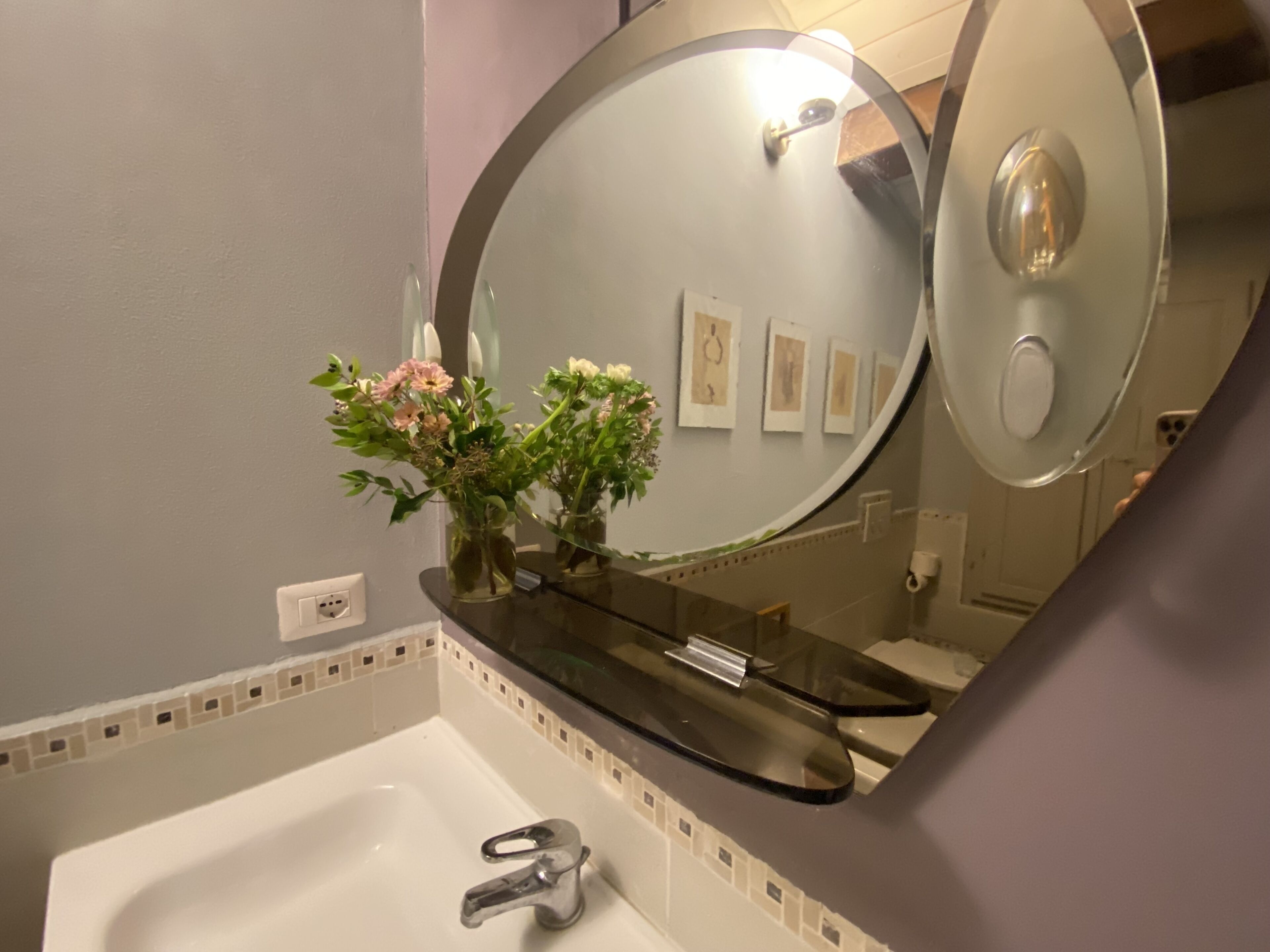 Un baño con lavamanos blanco, un espejo y un florero con flores.