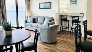 Una sala de estar con un sofá gris, una mesa de comedor redonda y dos sillones. Hay una zona de cocina con una barra y un cartel que dice "BEACH HOUSE".