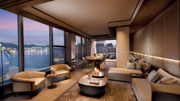Un salón moderno con un ventanal grande que ofrece vistas a un paisaje urbano y al agua. Hay un sofá, una mesa de centro y un comedor con una mesa y sillas.