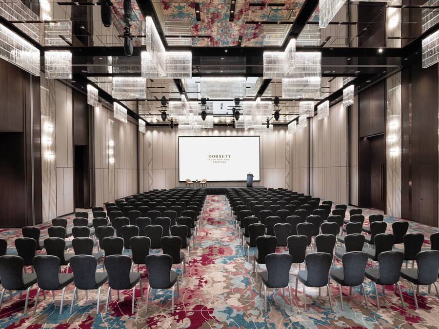 Un salón de conferencias con filas de sillas, una pantalla grande y una alfombra estampada con motivos florales.