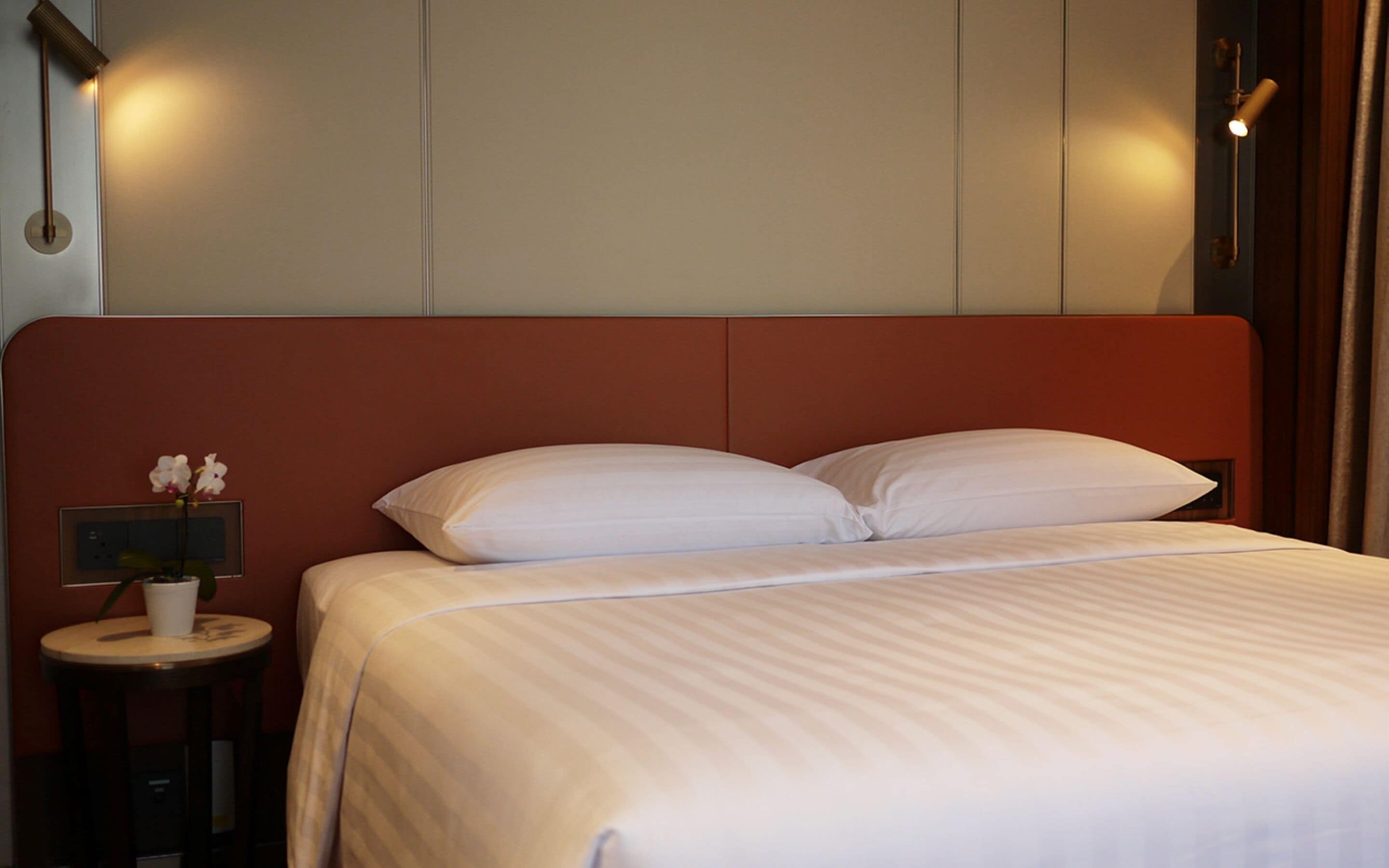 A neatly made bed with white linens, a small bedside table with a lamp, and a wall-mounted light fixture.