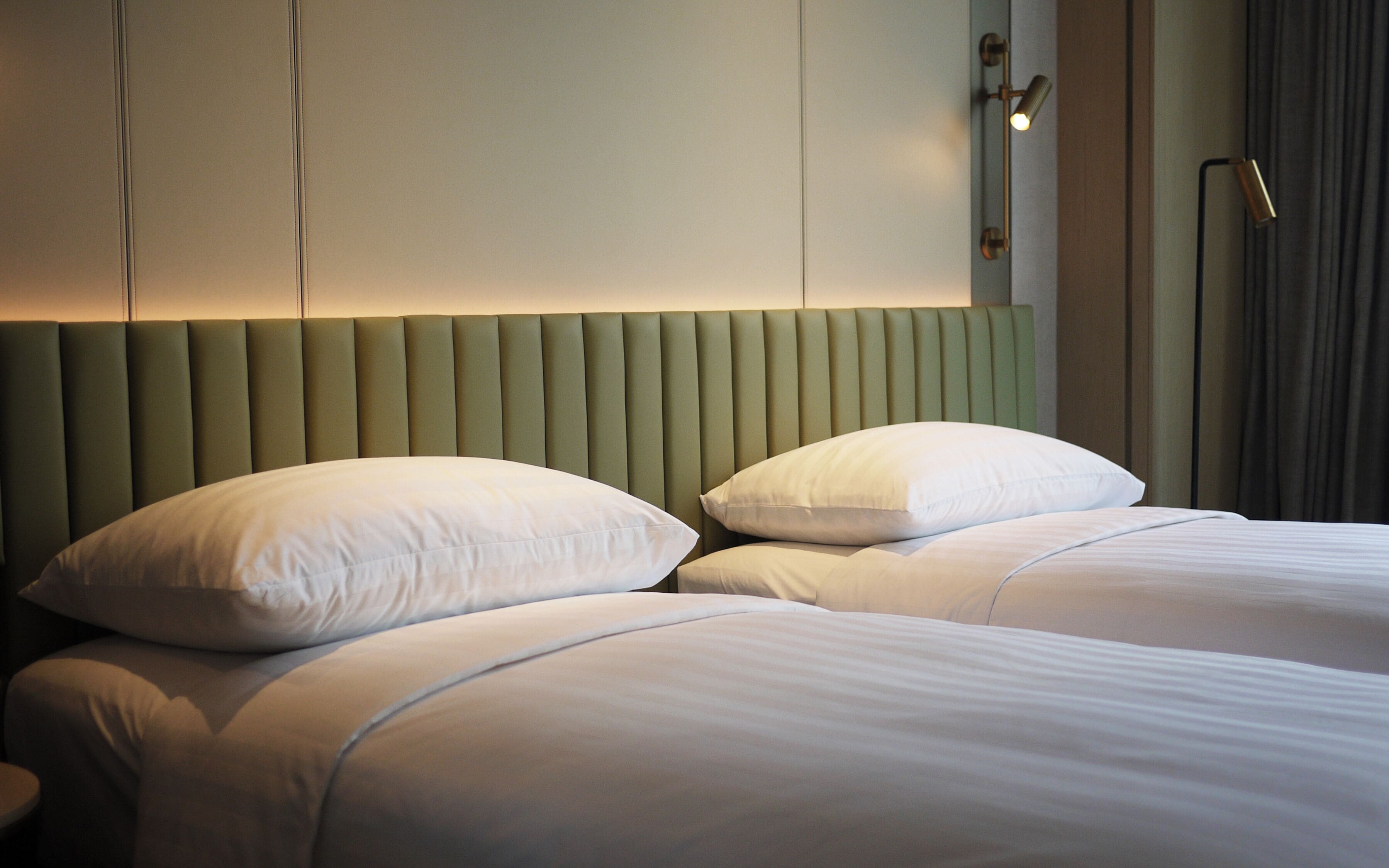 A neatly made bed with white linens and two pillows, a green upholstered headboard, and a bedside lamp.