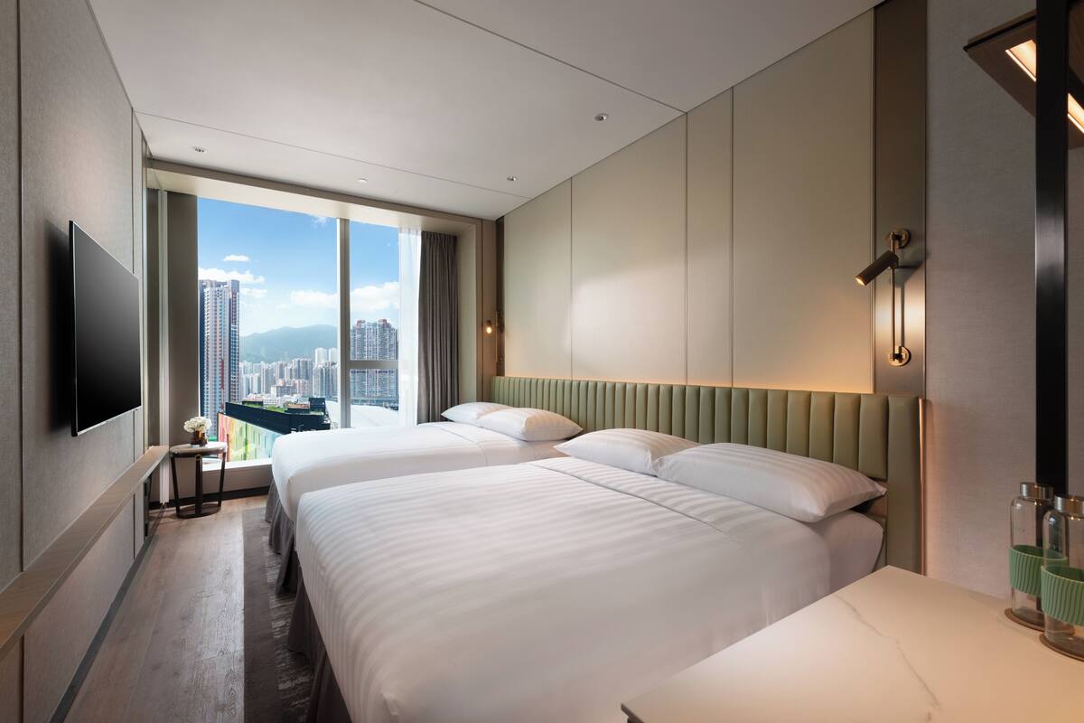 A modern hotel room with two beds, a flat-screen TV, a large window with a city view, and a nightstand with a lamp.