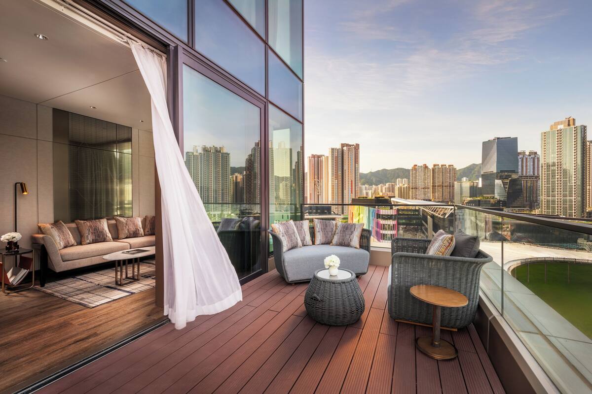 A modern balcony with a sofa, chairs, and a small table, overlooking a cityscape with high-rise buildings.