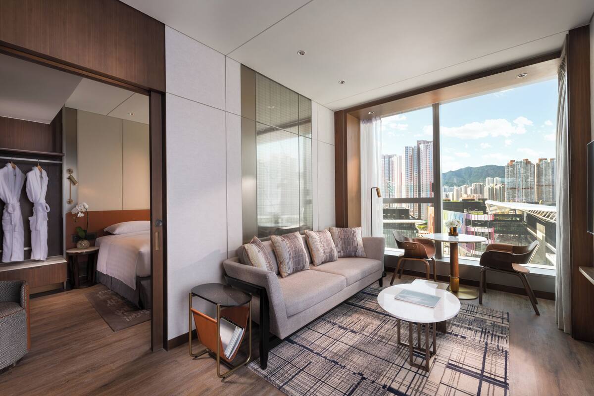 A modern hotel room with a large bed, a sofa, a dining area, and a view of the city.