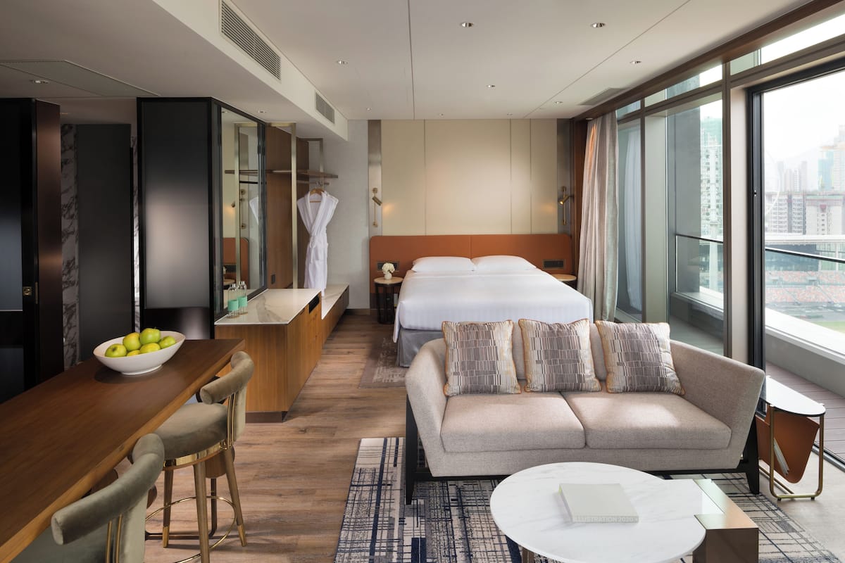 A modern hotel room with a sofa, a dining area with a bowl of fruit, a bed with a view, and a large window.