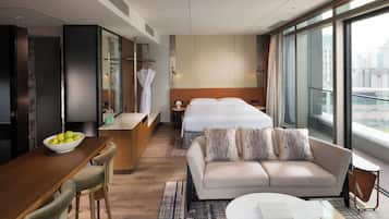 A modern hotel room with a sofa, a dining area with a bowl of fruit, a bed with a view, and a large window.
