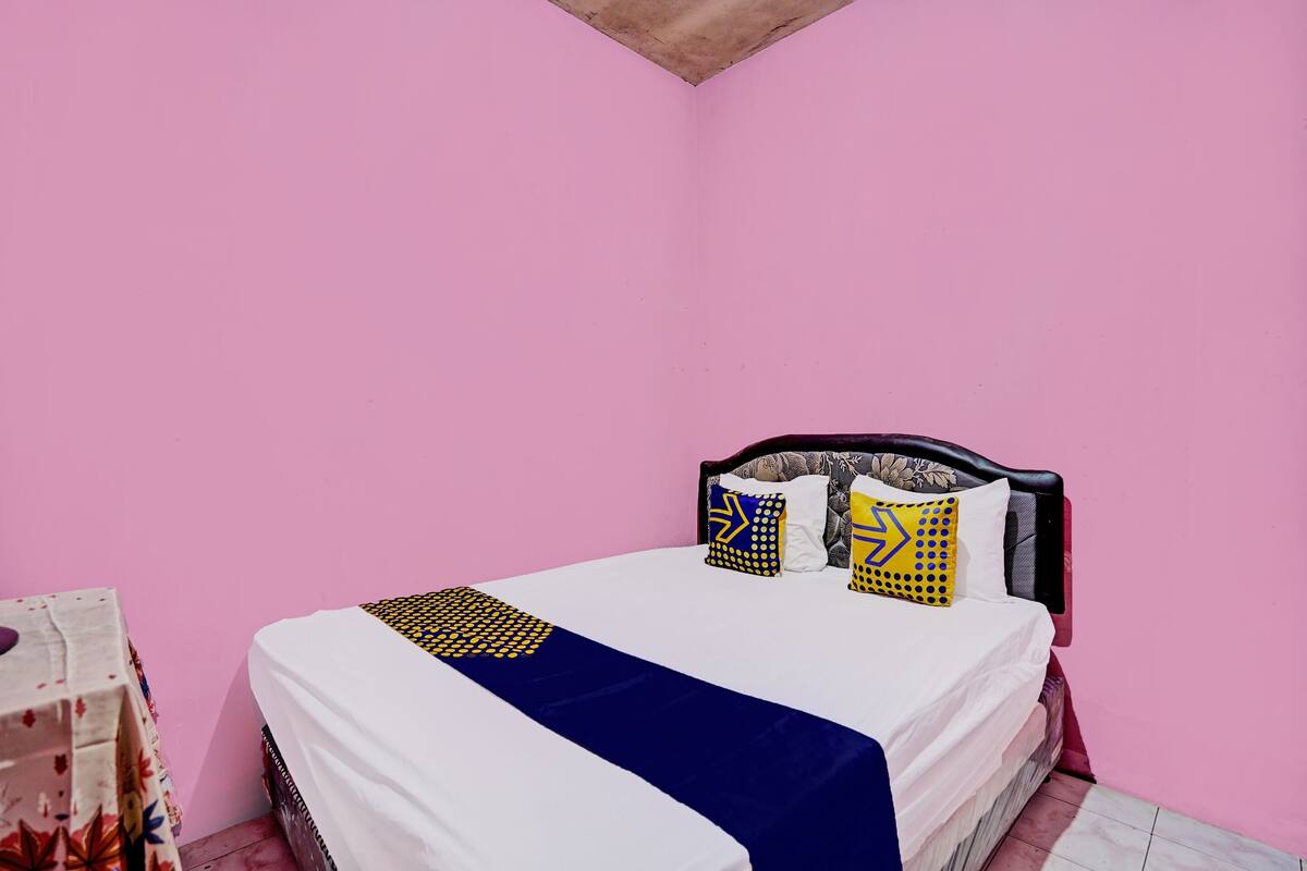 A bedroom with a bed against a pink wall, featuring a white mattress, a blue bedspread, and patterned pillows.
