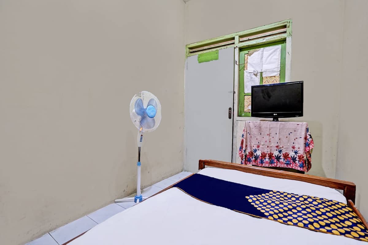 A room with a bed, a standing fan, a television on a table, and a window with a green frame.