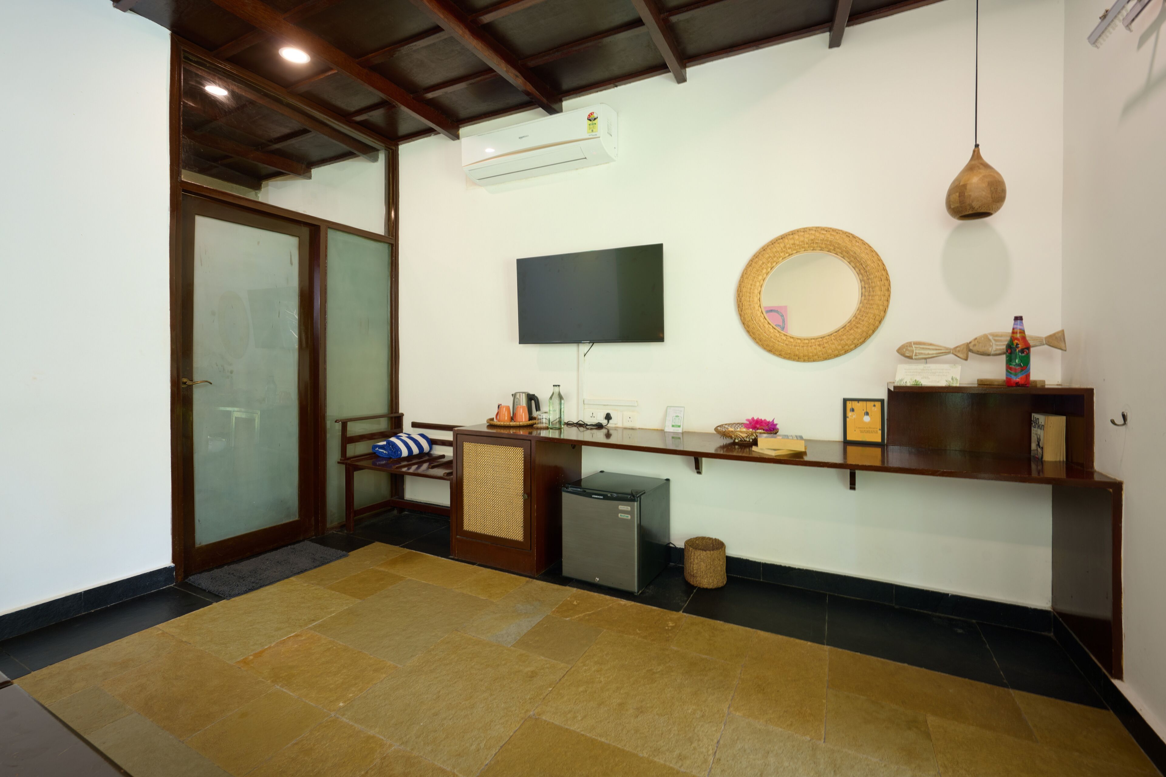 A compact living space with a sliding glass door, a flat-screen TV mounted on the wall, a woven basket, a small refrigerator, and a wooden shelf with various items.