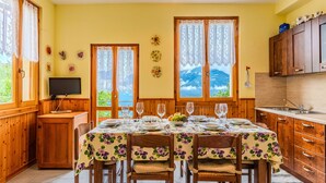A dining area with a table set for four, wooden cabinets, a kitchen area, and a view of mountains through large windows. - Casa Rita by Interhome (Valtellina)