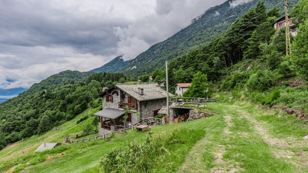 A rustic stone house with a wooden porch, surrounded by lush greenery and a mountain backdrop. - Casa Rita by Interhome (Valtellina)