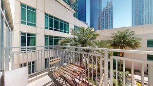 A balcony with wooden furniture and a white railing, overlooking a cityscape with tall buildings and palm trees. - 1 Bed Downtown| Walk to Dubai Mall & Burj Khalifa (Downtown Dubai)