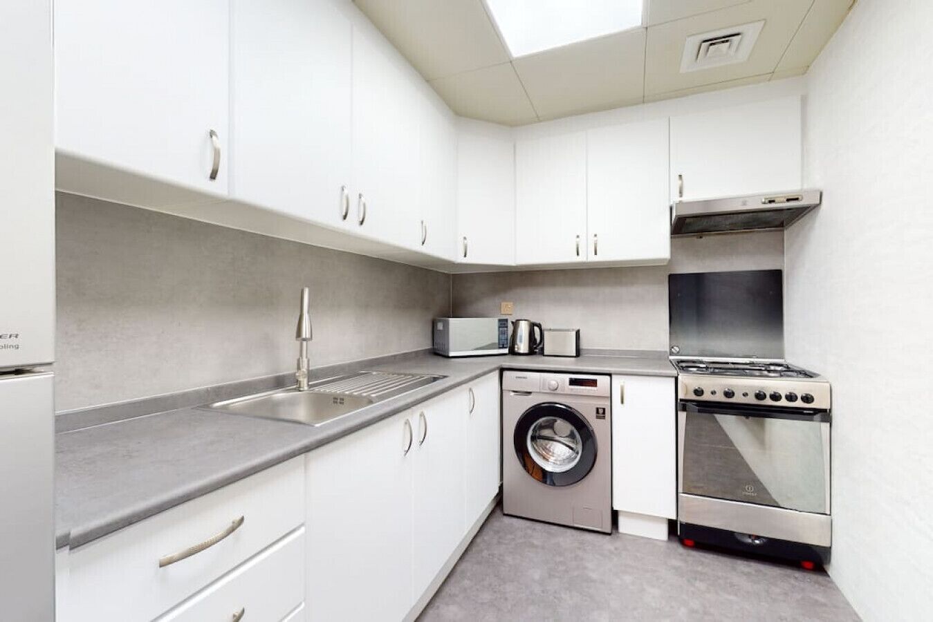 Cocina compacta con gabinetes blancos, un lavamanos de acero inoxidable, un microondas, una lavadora y una estufa a gas.
