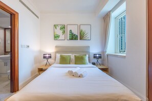 A hotel room with a bed, two bedside lamps, a window with curtains, and three framed pictures on the wall.