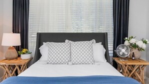 A neatly arranged bedroom with a bed, two bedside tables, a lamp, a fan, and a vase with flowers.
