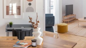 A modern living room with a wooden dining table, a sofa, a TV on a stand, and a large window. - Flawless 1BR Apartment in Athens (Menemeni)