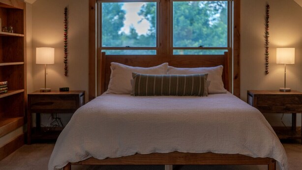 A bedroom with a wooden bed, two bedside tables, a nightstand, a bookshelf, and a window with a view of trees. - The Garden House in Livingston (Livingston)