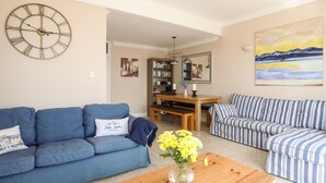 A living room with a blue sofa, a striped sofa, a wooden coffee table with a vase of flowers, a dining table with chairs, and a large wall clock. - 7 Min Y Traeth (Pwllheli)