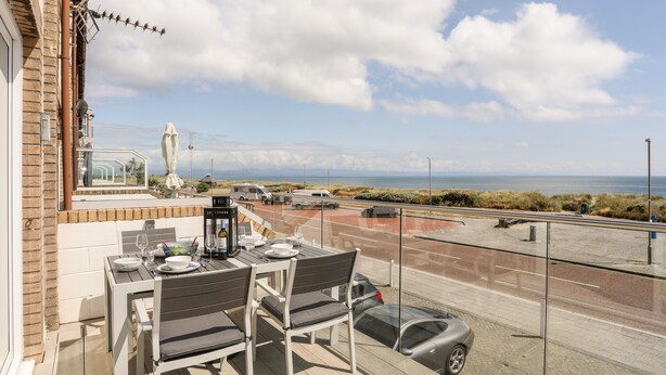 A balcony with a table set for dining, overlooking a coastal road and beach. - 7 Min Y Traeth (Pwllheli)