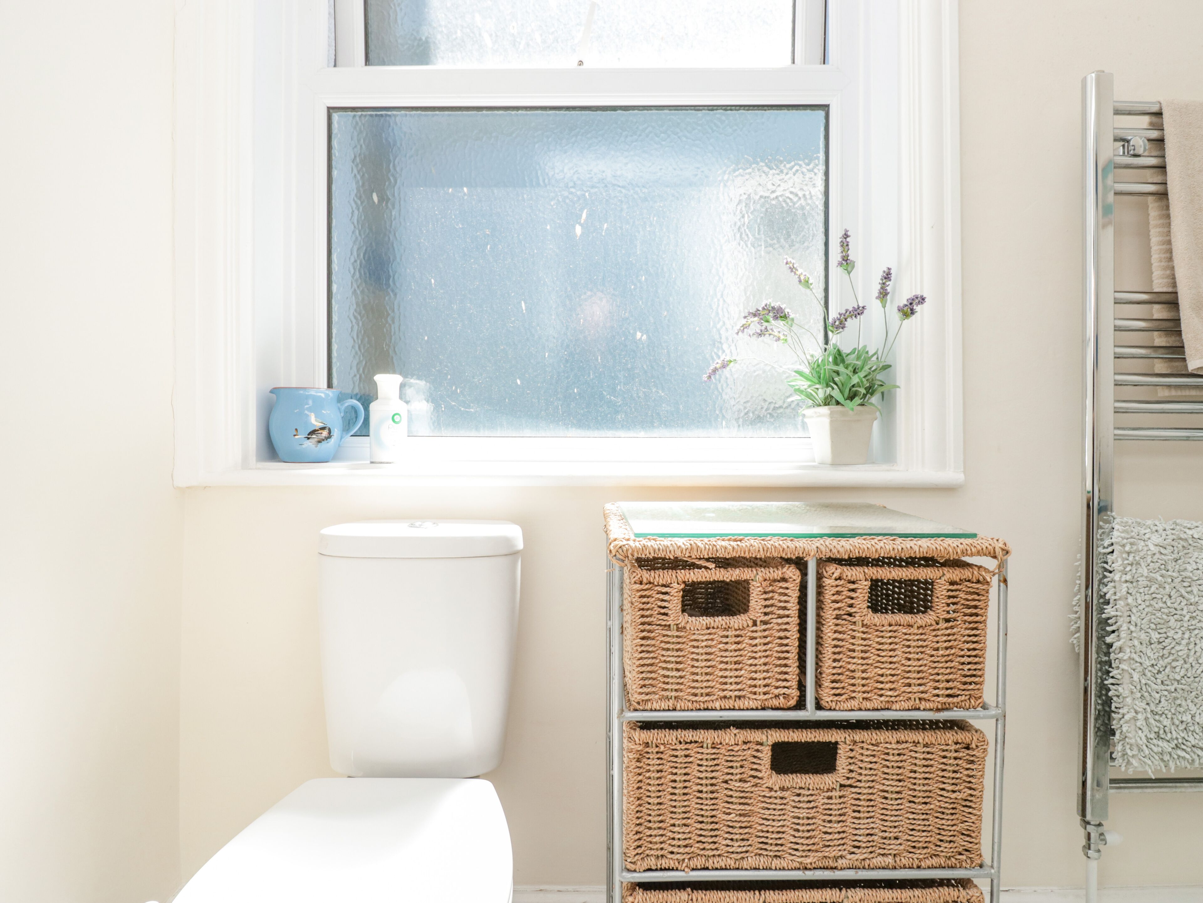A bathroom with a toilet, a window with a frosted glass, a blue mug, a white bottle, and wicker baskets.