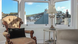 A room with a large bay window, a plush armchair, a small table with a lamp, and a view of a coastal town. - 29b Lower Street (Dartmouth)