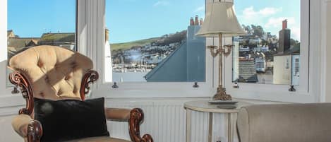A room with a large bay window, a plush armchair, a small table with a lamp, and a view of a coastal town.