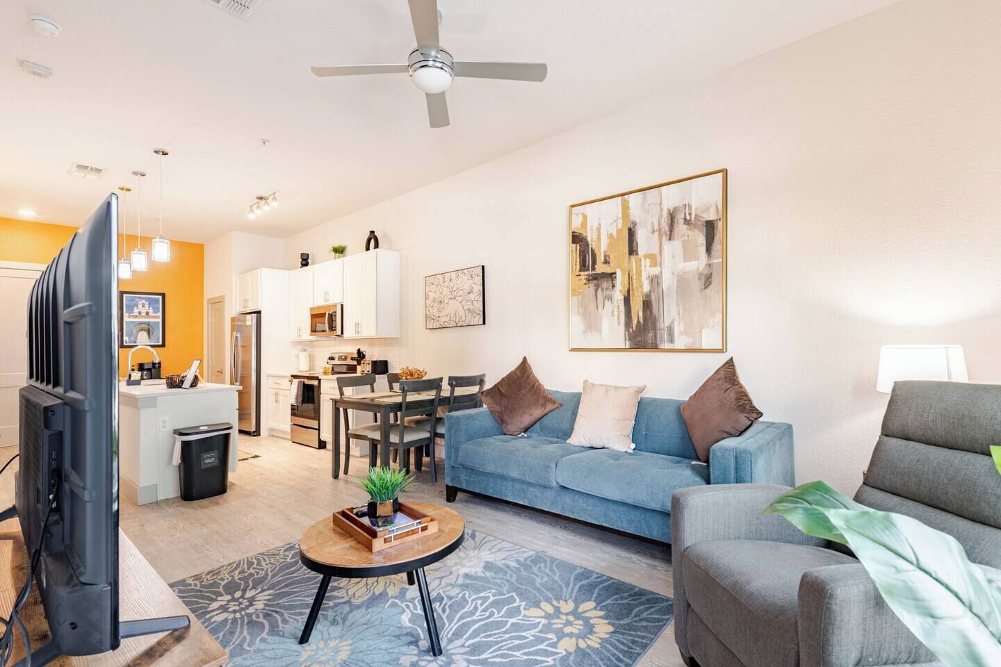 Un salon moderne avec un canapé, un fauteuil et une table basse. On aperçoit une télévision, un ventilateur de plafond et une cuisine au fond.