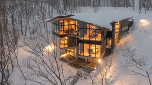 A modern, multi-level cabin with large glass windows, surrounded by a snowy forest. - Hanaridge Residence 11 (Kutchan)
