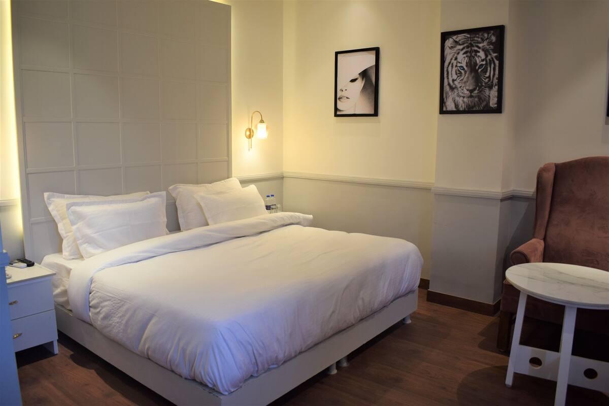 A neatly made bed with white linens, a bedside table with a lamp, a chair, and two framed pictures on the wall.