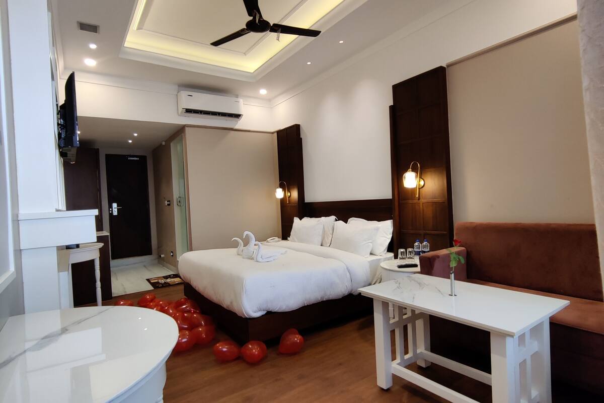 A modern hotel room with a large bed, a ceiling fan, an air conditioning unit, a white bedside table, a brown sofa, and a small round table.